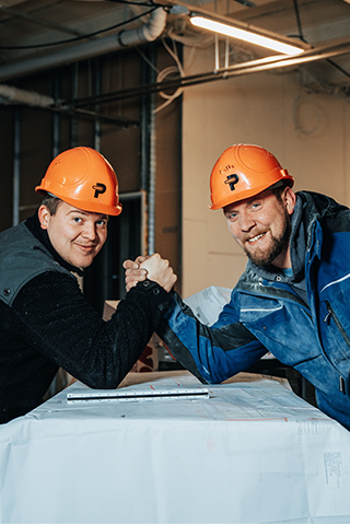 In der Mitte stehen zwei POHL-Mitarbeiter mit orangefarbenen Helmen einander zugewandt und fassen sich über einer Tischkante an den Händen. Links und rechts im Bild sind helle Innenwände und Metallprofile eines Rohbaus. Im Hintergrund stehen weitere Personen mit Helmen. Vorne im Bild liegen Pläne und ein Lineal auf dem Tisch.