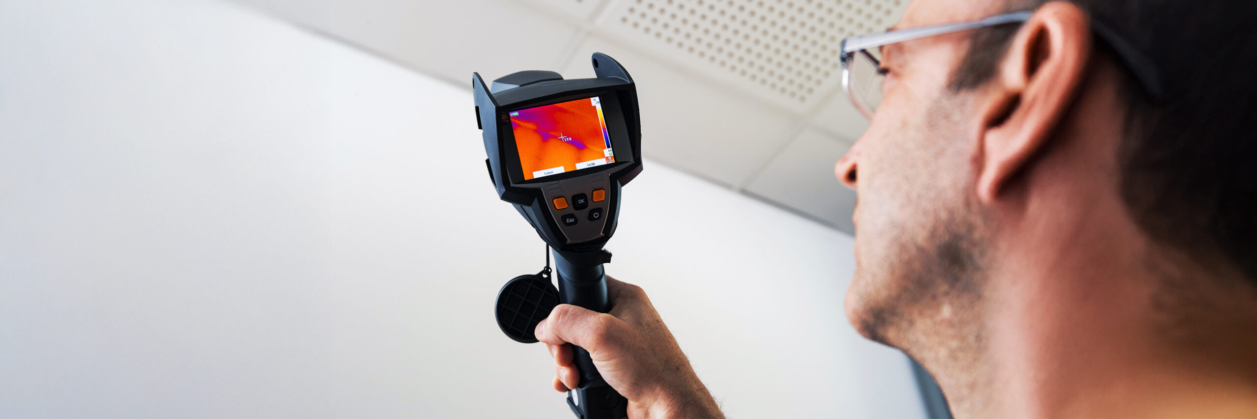 Ein Mann hält ein Messgerät mit Bildschirm, auf dem farbige Wärmebilder angezeigt werden. Er prüft damit die Isolierung eines Innenraums.