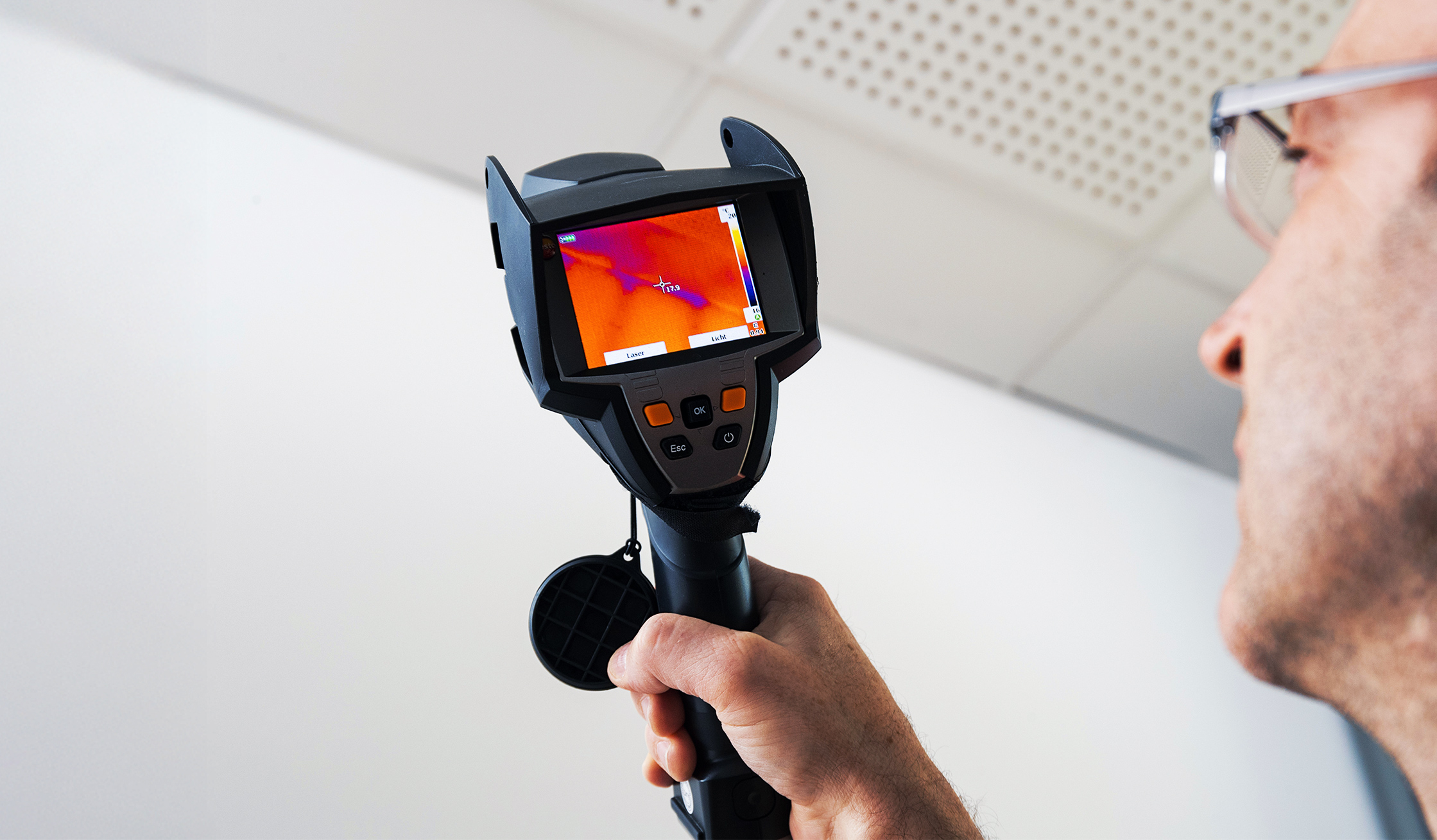 Ein Mann hält ein Messgerät mit Bildschirm, auf dem farbige Wärmebilder angezeigt werden. Er prüft damit die Isolierung eines Innenraums.