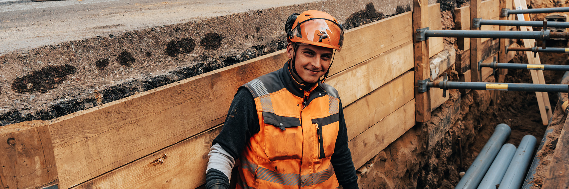 In der Mitte steht ein POHL-Mitarbeiter in orangefarbener Weste und Helm in einem schmalen, seitlich mit Holzbrettern abgestützten Graben. Rechts im Bild halten graue Querstreben die Holzseiten. Links oben im Bild verläuft der Rand einer Asphaltfläche. Im Hintergrund liegen mehrere blaue Rohre nebeneinander im Graben.