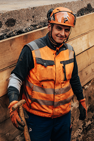 In der Mitte steht ein POHL-Mitarbeiter in orangefarbener Weste und Helm in einem schmalen, seitlich mit Holzbrettern abgestützten Graben. Rechts im Bild halten graue Querstreben die Holzseiten. Links oben im Bild verläuft der Rand einer Asphaltfläche. Im Hintergrund liegen mehrere blaue Rohre nebeneinander im Graben.