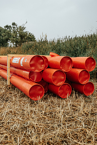 Vorne im Bild liegen mehrere orangefarbene Rohre in zwei Reihen auf Holzböcken; rechts am Rand wächst hohes Schilf, links verläuft ein Stoppelfeld mit einem einzelnen Baum im Hintergrund.