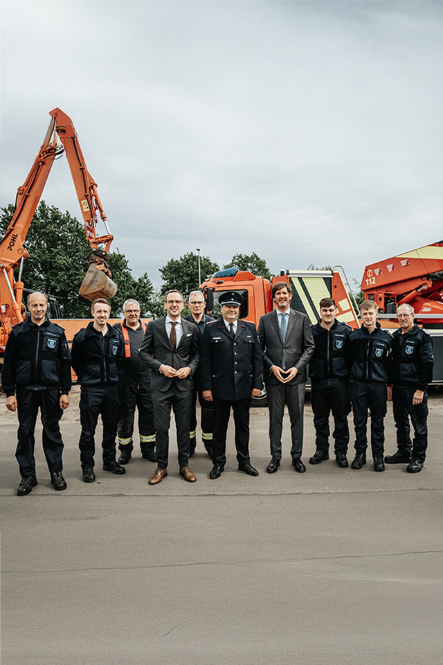 Gruppenfoto vor Einsatzfahrzeugen mit Reihen von Einsatzkräften in dunklen Uniformen und zwei Personen im Anzug in der Mitte, links im Hintergrund ein Baggerarm mit Pohl Schriftzug, rechts eine Feuerwehr-Drehleiter, grauer Himmel.