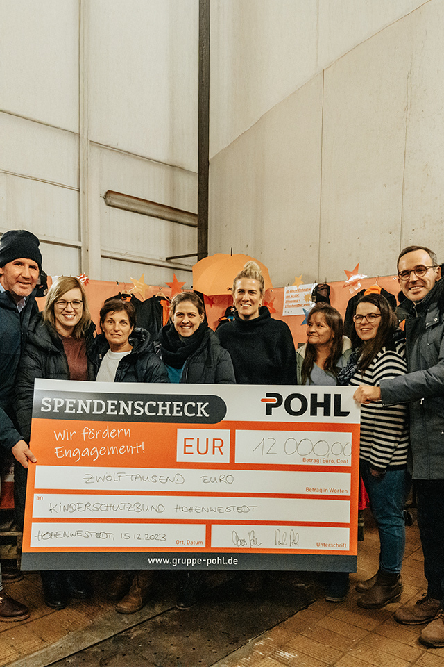 Innenaufnahme eines Spendenevents mit einer Gruppe Frauen und Männer, die gemeinsam einen übergroßen Scheck quer halten; auf dem Scheck Pohl Schriftzug, im Hintergrund Ständer mit Kleidung und orange Dekoration.