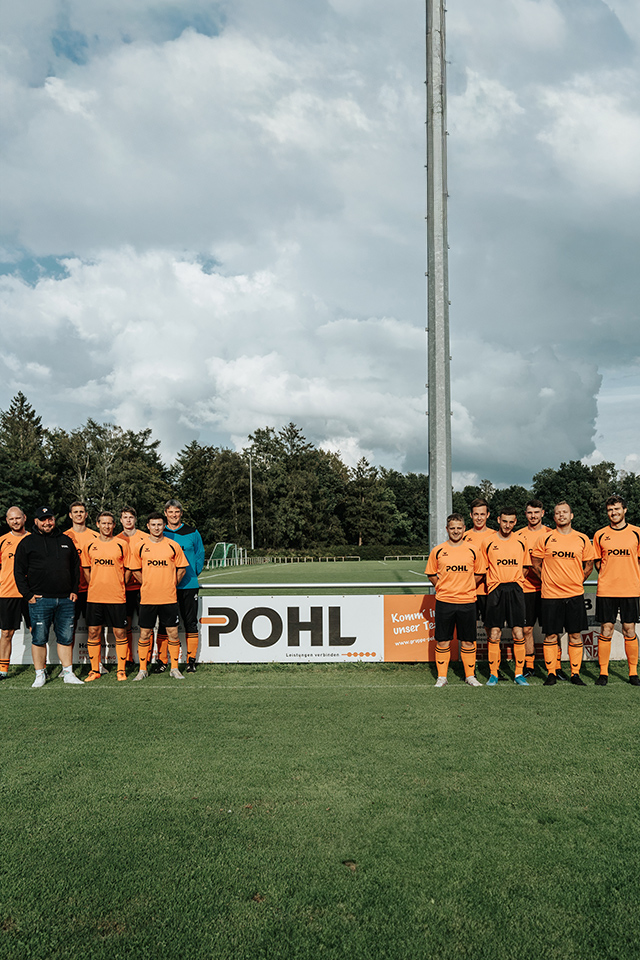 Fußballteam am Spielfeldrand: eine Reihe Spieler in orangefarbenen Trikots steht an der Seitenbande; auf Bande und Trikots Pohl Schriftzug, grüner Rasen und Bäume dahinter.