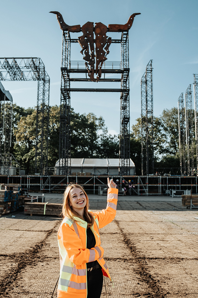 Offenes Festival-/Baustellengelände bei Sonne: vorne mittig eine Frau in Warnjacke zeigt lächelnd auf die hohe Bühnenkonstruktion mit Stierschädel-Symbol; Gerüsttürme im Hintergrund, Boden mit Fahrplatten.