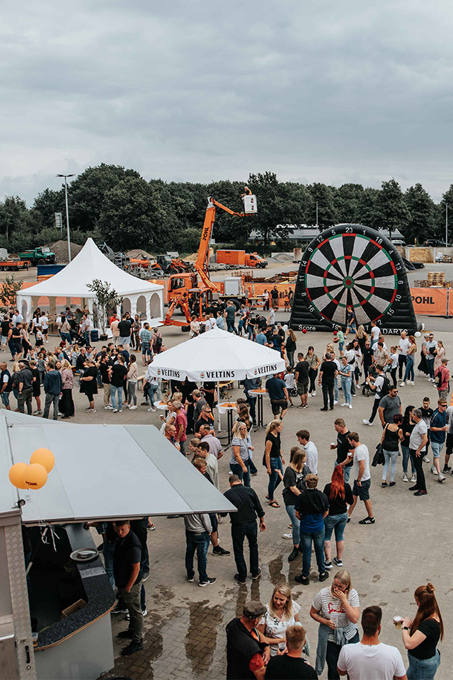 Weitwinkelaufnahme eines Firmenevents auf dem Betriebshof: vorne links Food-Standdächer, mittig mehrere weiße Pagodenzelte und Menschen in Gruppen, rechts Hüpfburg und Spielfläche, mittig hinten eine riesige aufblasbare Dartscheibe; orange Absperrzäune mit POHL Schriftzug im Hintergrund, Bau- und Logistikfahrzeuge dahinter, bewölkter Himmel.