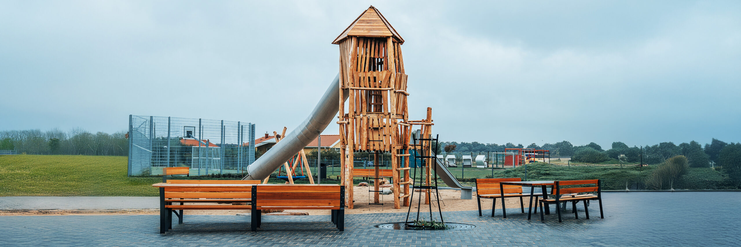 Referenz Attraktivierung Kurpark Friedrichskoog: Vorne zwei Holzbänke, dahinter mittig ein hoher Holzturm mit langer silberner Röhrenrutsche nach links; rechts daneben eine zweite, gerade Rutsche; im Hintergrund Zäune und Wiesen.