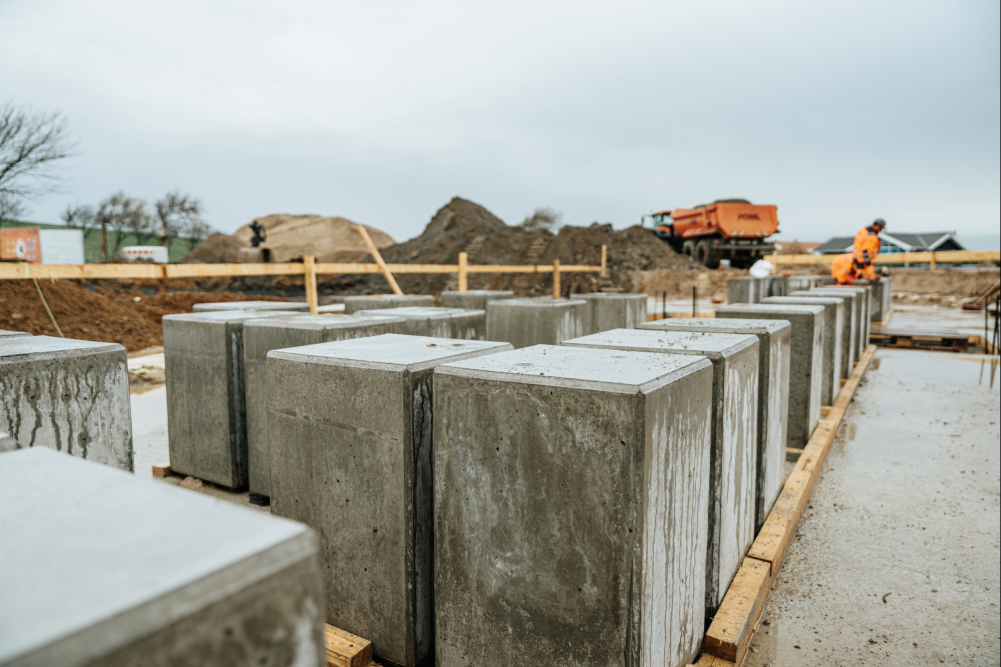Mehrere massive Betonblöcke stehen in gleichmäßigen Reihen auf einer Baustelle. Im Hintergrund sind Erdhügel, ein orangefarbener LKW und zwei Arbeiter in orangefarbener Schutzkleidung zu sehen. Der Boden ist feucht, und der Himmel ist grau.