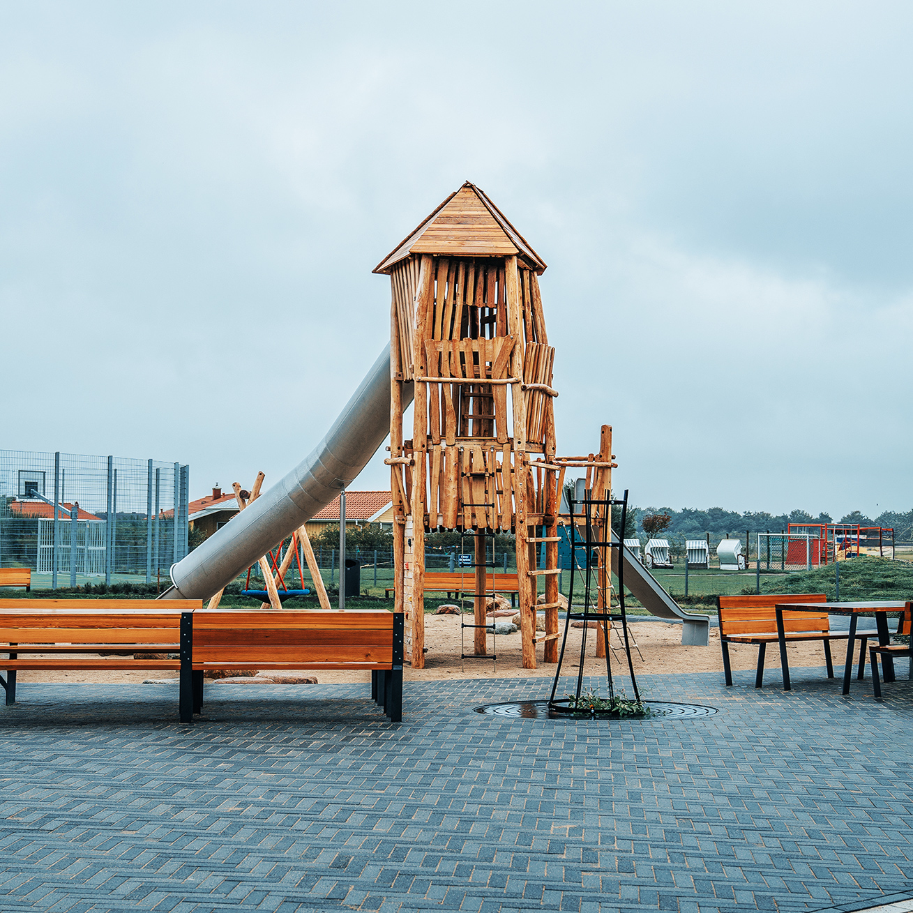 Referenz Attraktivierung Kurpark Friedrichskoog: Vorne zwei Holzbänke, dahinter mittig ein hoher Holzturm mit langer silberner Röhrenrutsche nach links; rechts daneben eine zweite, gerade Rutsche; im Hintergrund Zäune und Wiesen.