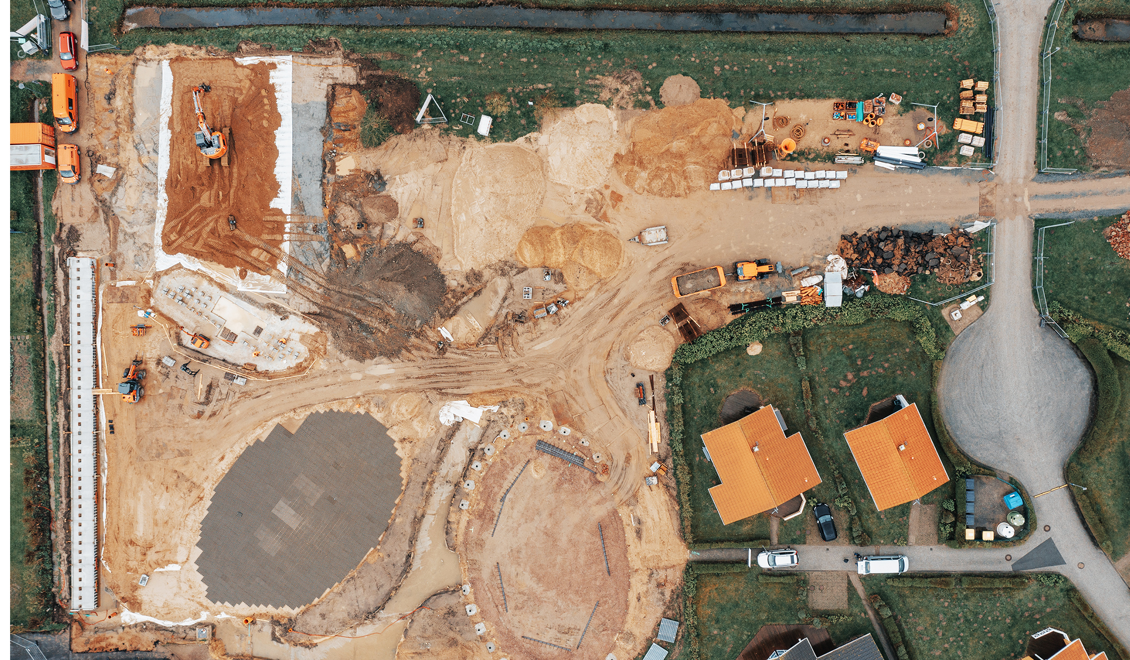 Referenz Attraktivierung Kurpark Friedrichskoog: Senkrechte Luftaufnahme einer Baustelle: links arbeitet ein orangefarbener POHL-Bagger auf einer großen Sandfläche; rechts kreuzen sich zwei Baustellenwege; oben und unten schmale Wassergräben; mehrere Materialhaufen und weitere orange POHL-Fahrzeuge.