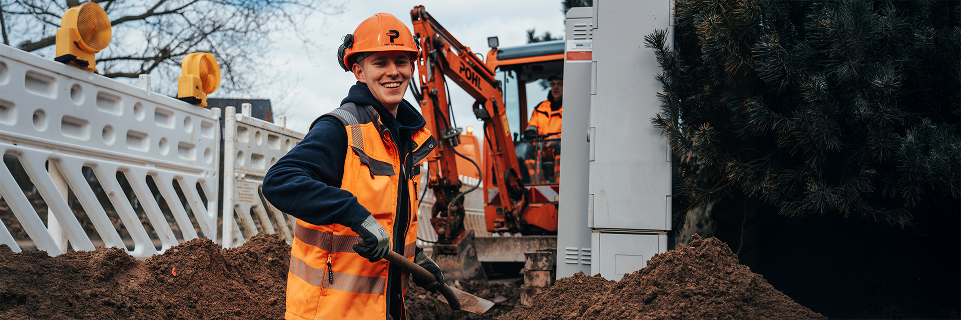 Vorne im Bild liegt ein aufgeschütteter Erdhaufen. Links im Bild steht ein POHL-Mitarbeiter mit orangefarbenem Helm und orangefarbener Weste mit einer Schaufel in der Hand. In der Mitte und nach rechts versetzt steht eine orangefarbene POHL-Maschine hinter weißen Absperrelementen. Rechts am Rand steht ein grauer Kasten neben dunklen Nadelbäumen. Im Hintergrund sind kahle Äste und weitere Absperrungen zu sehen.