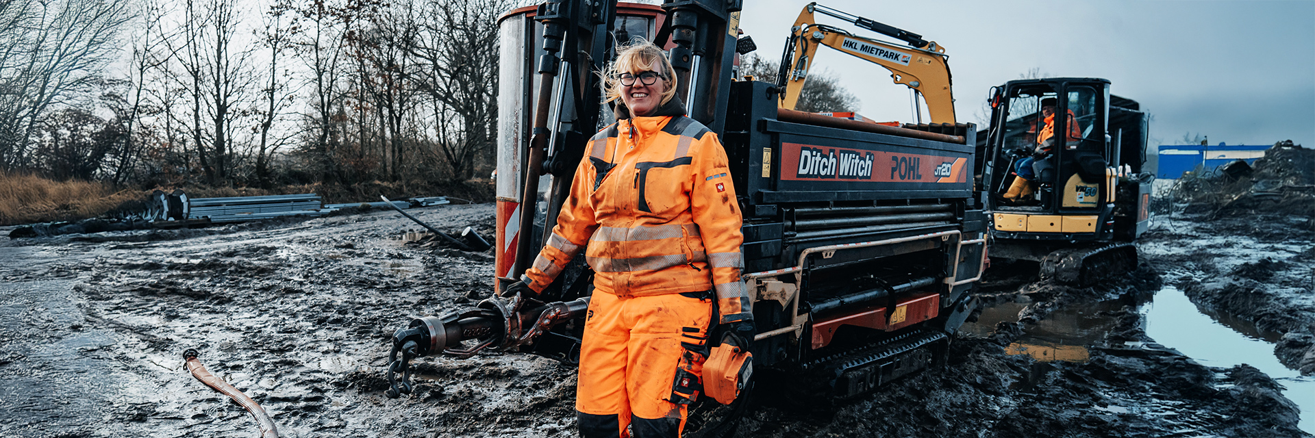 In der Mitte steht eine POHL-Mitarbeiterin in orangefarbener Arbeitskleidung neben einer schwarzen Maschine mit den Aufdrucken „Ditch Witch“ und „POHL“. Rechts im Bild steht eine gelbe Kettenmaschine mit einer Person in der Kabine. Links im Bild liegen Wasserpfützen auf schlammigem Boden. Im Hintergrund stehen kahle Bäume.