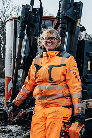 In der Mitte steht eine POHL-Mitarbeiterin in orangefarbener Arbeitskleidung neben einer schwarzen Maschine mit den Aufdrucken „Ditch Witch“ und „POHL“. Rechts im Bild steht eine gelbe Kettenmaschine mit einer Person in der Kabine. Links im Bild liegen Wasserpfützen auf schlammigem Boden. Im Hintergrund stehen kahle Bäume.