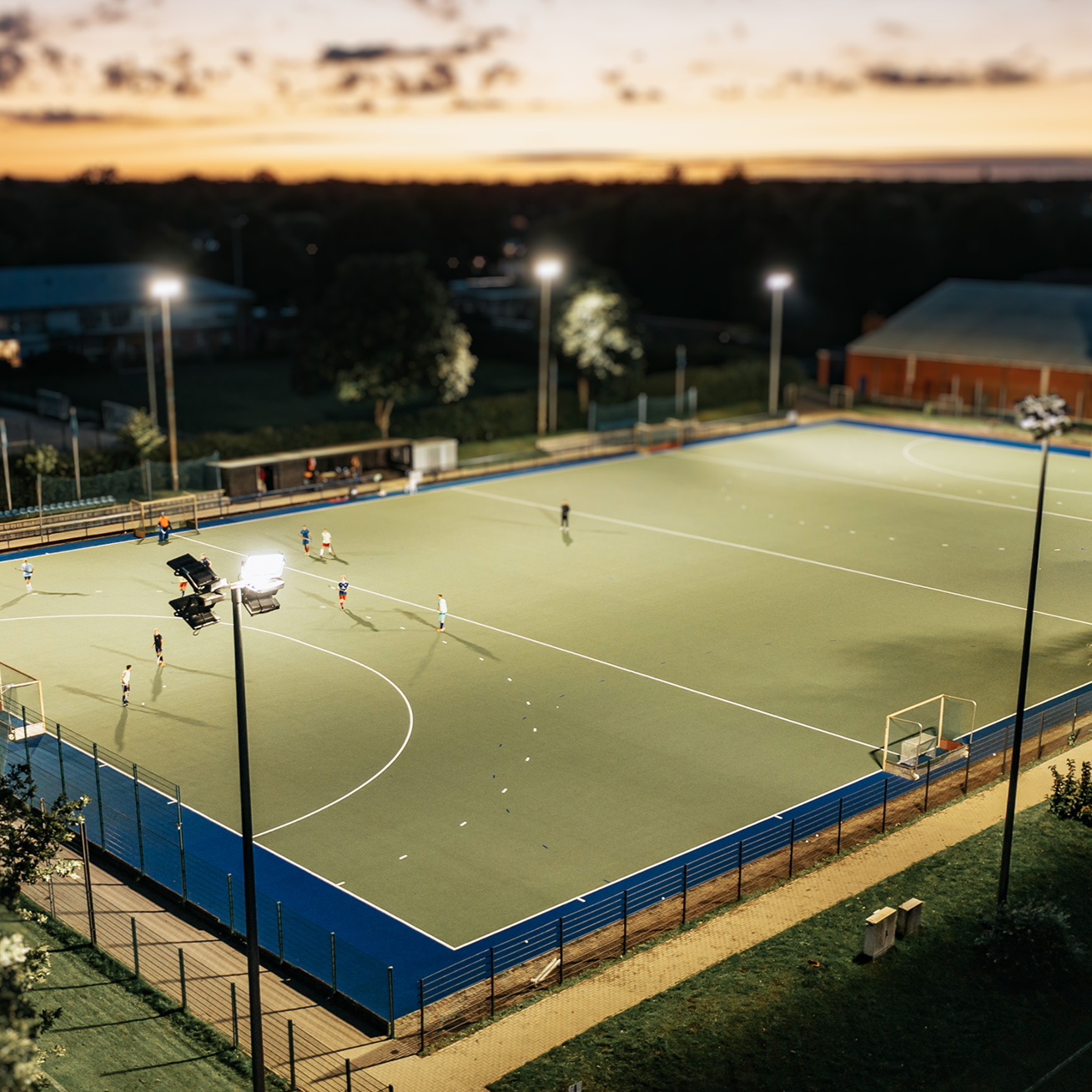 Referenz Flutlicht HSV Hamburg: Schräger Blick von oben auf ein hell beleuchtetes Spielfeld in der Dämmerung. In der Mitte und an den Ecken stehen hohe Masten; links vorne strahlt ein Flutlicht besonders hell auf den Platz. Rund um den Platz verläuft ein Zaun; im Hintergrund liegen Gebäude und Bäume im Dunkeln.