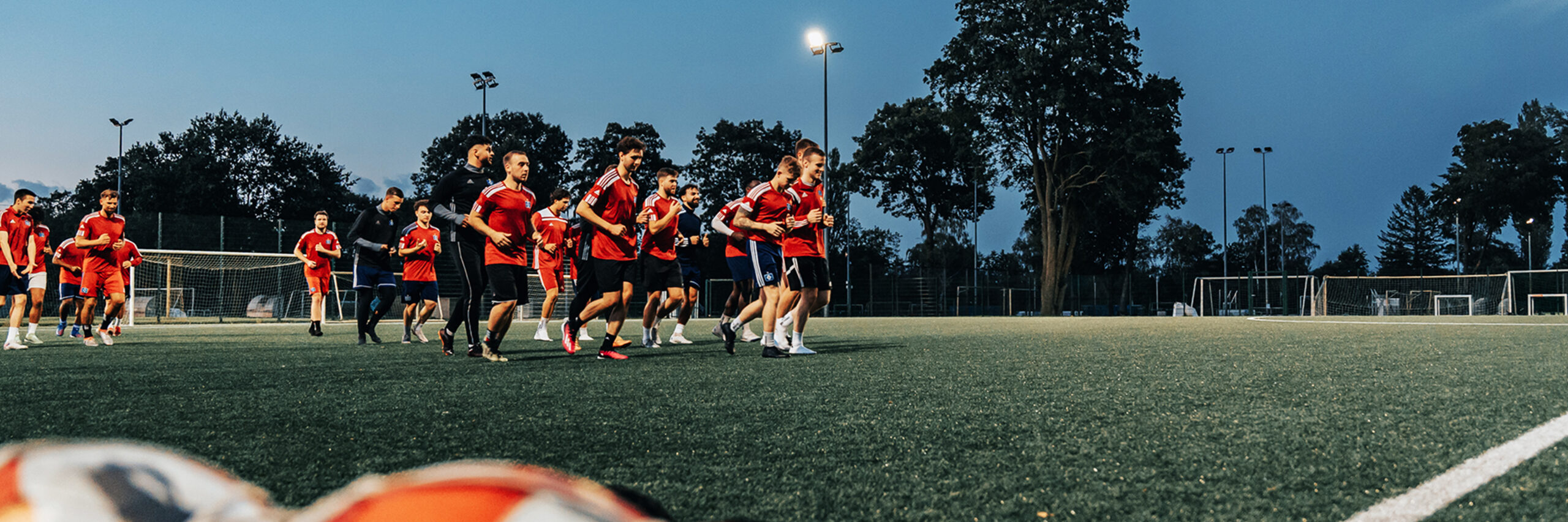 Referenz Flutlicht HSV Hamburg: Unten im Vordergrund liegen unscharf mehrere Fußbälle in Netzen. In der Mitte laufen Spieler in roten Trikots über das beleuchtete Spielfeld; links und rechts stehen Tore. Im Hintergrund ragen Masten mit eingeschaltetem Licht vor dem dunkler werdenden Himmel.