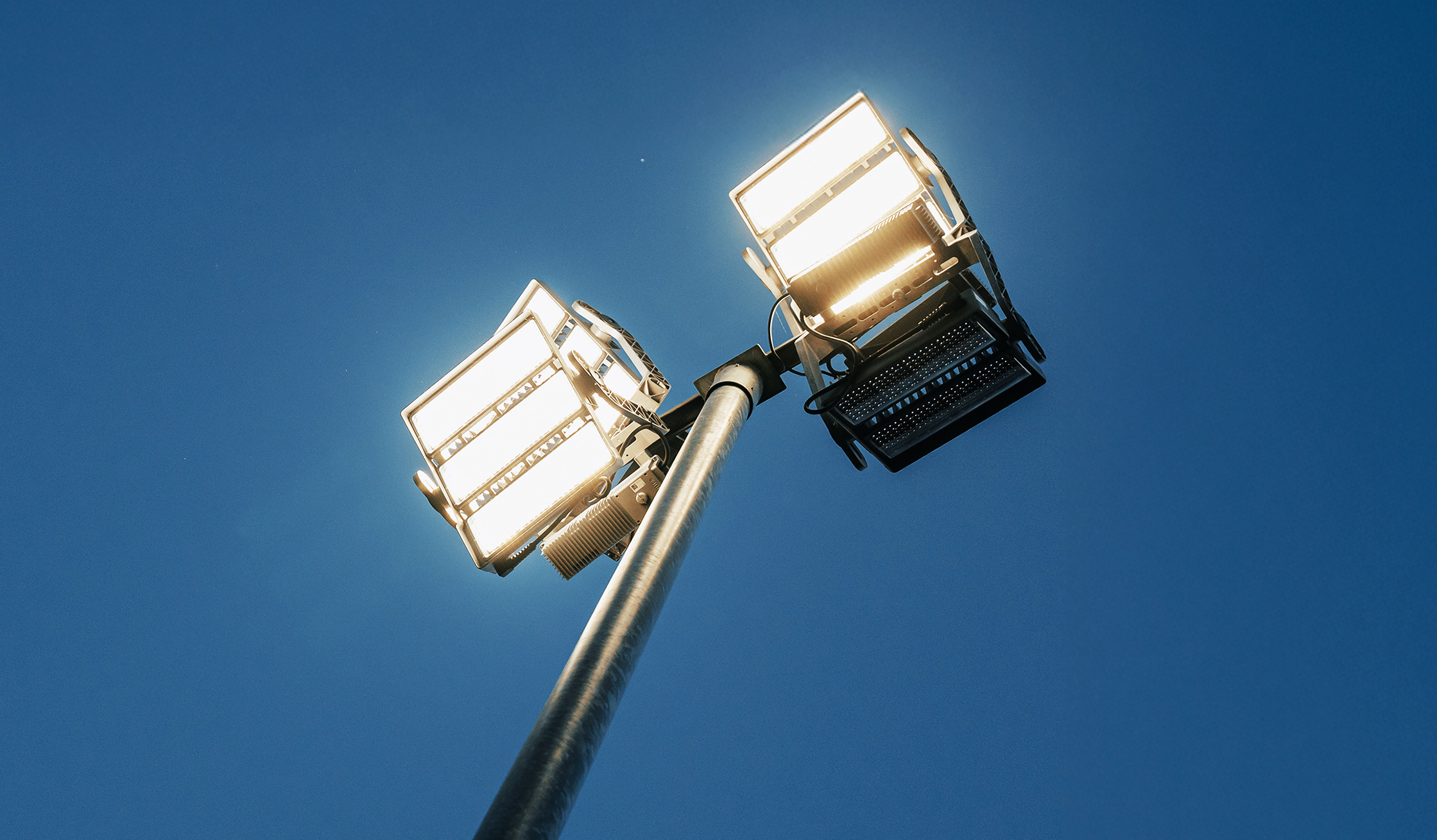 Referenz Flutlicht HSV Hamburg: Von unten gesehen leuchten oben im Bild zwei große Flutlichtköpfe an einem Mast. Am Gehäuse sind Halterungen und Kabel zu erkennen; der Hintergrund zeigt klaren Himmel ohne weitere Elemente.