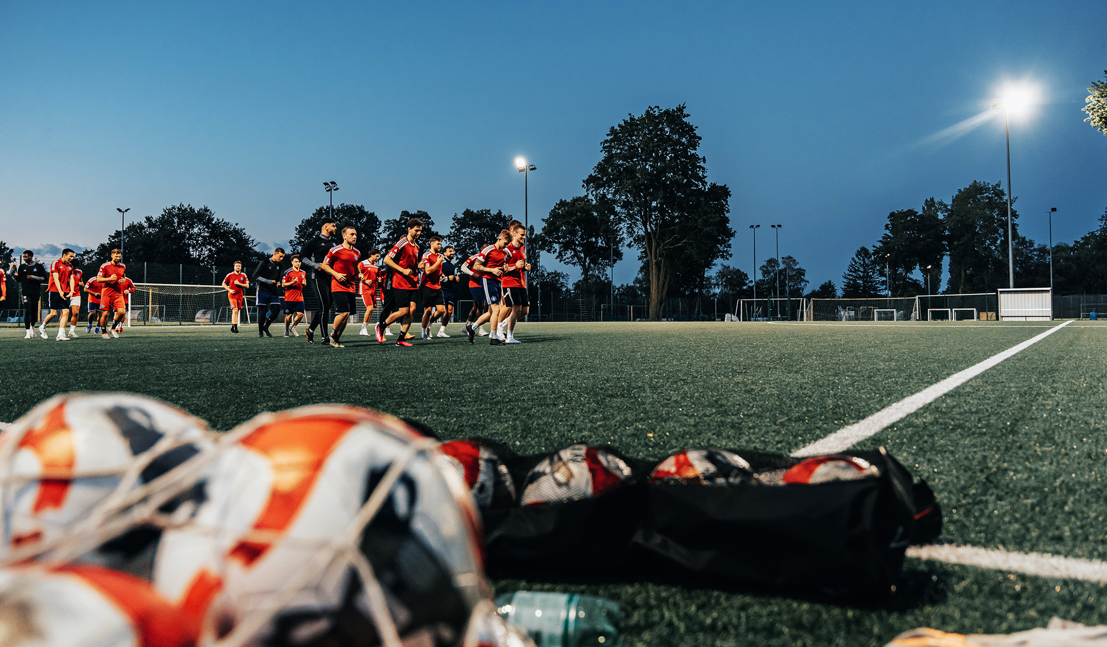 Referenz Flutlicht HSV Hamburg: Unten im Vordergrund liegen unscharf mehrere Fußbälle in Netzen. In der Mitte laufen Spieler in roten Trikots über das beleuchtete Spielfeld; links und rechts stehen Tore. Im Hintergrund ragen Masten mit eingeschaltetem Licht vor dem dunkler werdenden Himmel.