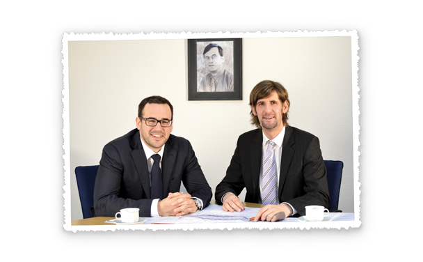 Zwei Männer in Anzügen sitzen am Schreibtisch und blicken in die Kamera, über ihnen hängt ein gerahmtes Porträt.