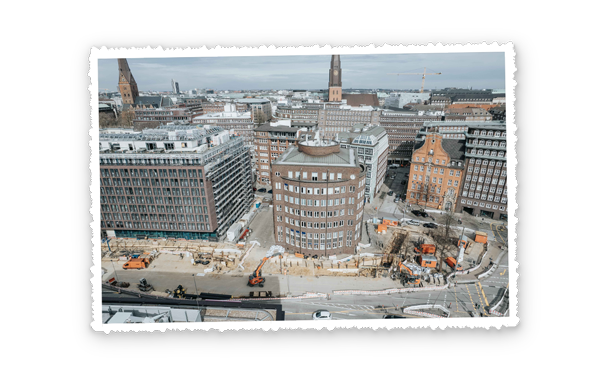 Blick aus der Höhe über eine große Stadtkreuzung, im Vordergrund eine Baustelle mit mehreren orangefarbenen Fahrzeugen und Geräten, darum Büro- und Wohngebäude.