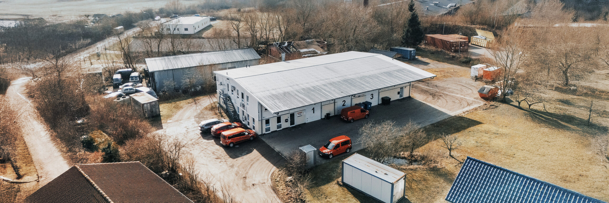 Standort Ahrensbök, Luftaufnahme: In der Mitte liegt dasselbe Gebäude mit den Toren 1 bis 3; davor und links parken mehrere orange POHL-Fahrzeuge. Rechts am Rand stehen Container und Bäume; hinten schließen Felder an.