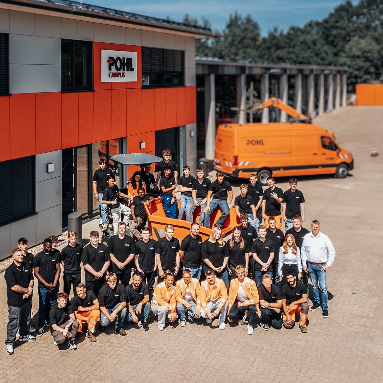 Großes Gruppenfoto von POHL-Auszubildenden vor dem Gebäude „POHL Campus“; viele in schwarzen Shirts, einige in oranger Arbeitskleidung, rechts im Hof ein orangefarbener Transporter.