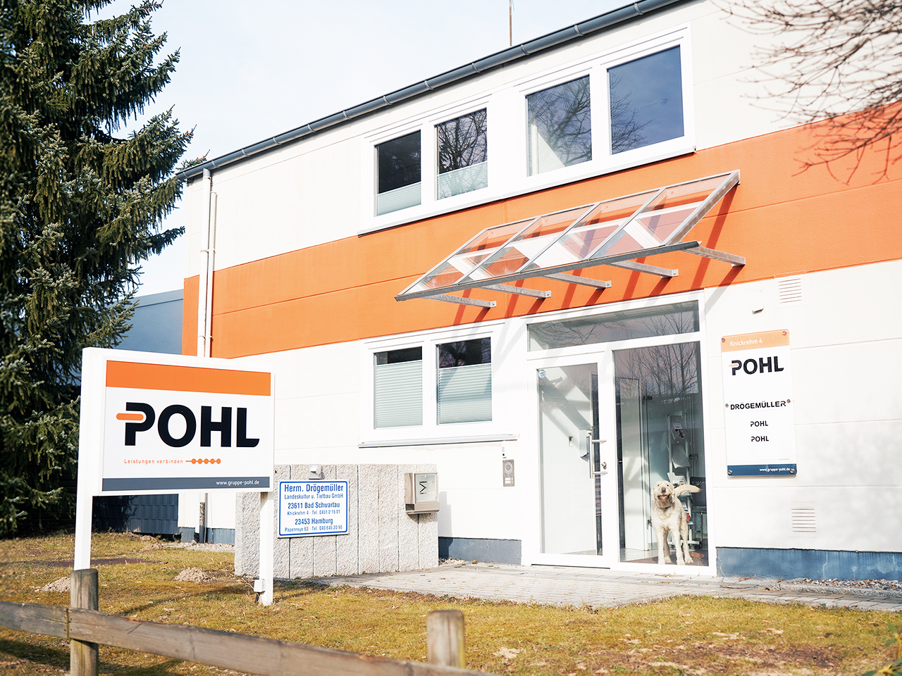 Standort Bad Schwartau: In der Mitte befindet sich der Eingang eines zweigeschossigen Gebäudes mit orangefarbener Fassadenlinie; rechts neben der Tür hängt ein POHL-Schild. Links vorne steht ein freistehendes POHL-Schild; hinter der Glastür steht ein heller Hund.