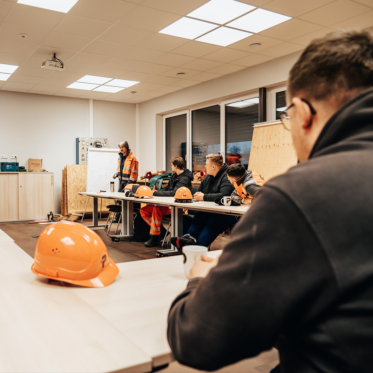 Bild in den Räumen des POHL-Campus. In der Mitte steht ein POHL-Mitarbeiter in orange Jacke vor einem Flipchart und erklärt etwas. Vorne auf dem Tisch liegen mehrere orange Schutzhelme; links an der Wand stehen Schränke, rechts geben Fenster den Blick nach draußen frei.