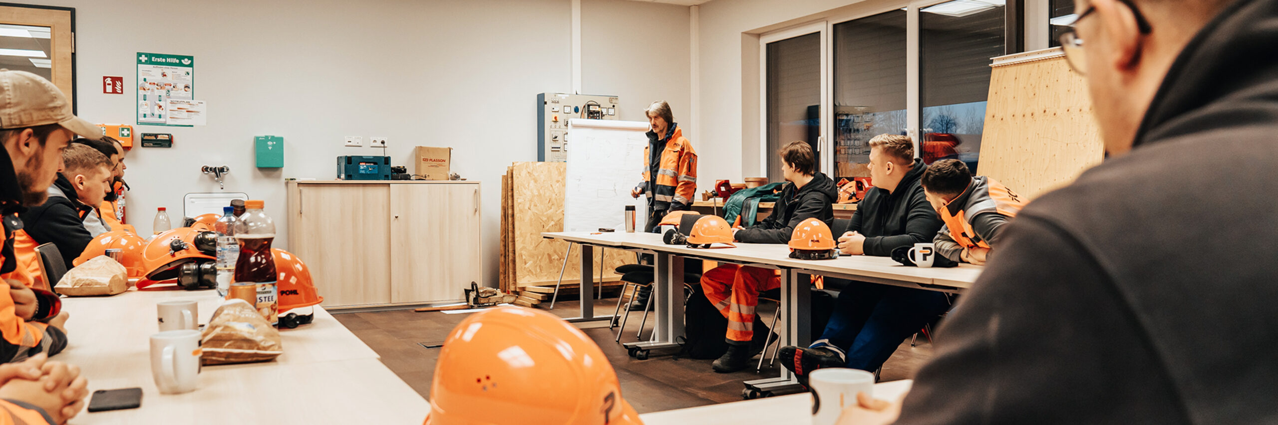 In der Mitte steht ein POHL-Mitarbeiter in orange Jacke vor einem Flipchart und erklärt etwas. Vorne auf dem Tisch liegen mehrere orange Schutzhelme; links an der Wand stehen Schränke, rechts geben Fenster den Blick nach draußen frei.