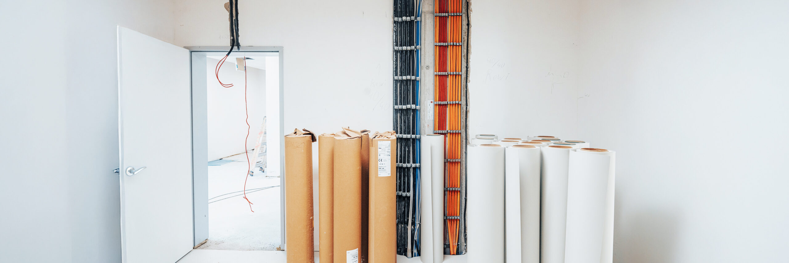 Innenaufnahme einer Baustelle: Kabelstränge verlaufen geordnet an einer Wand, daneben stehen Rollen mit Baumaterialien.
