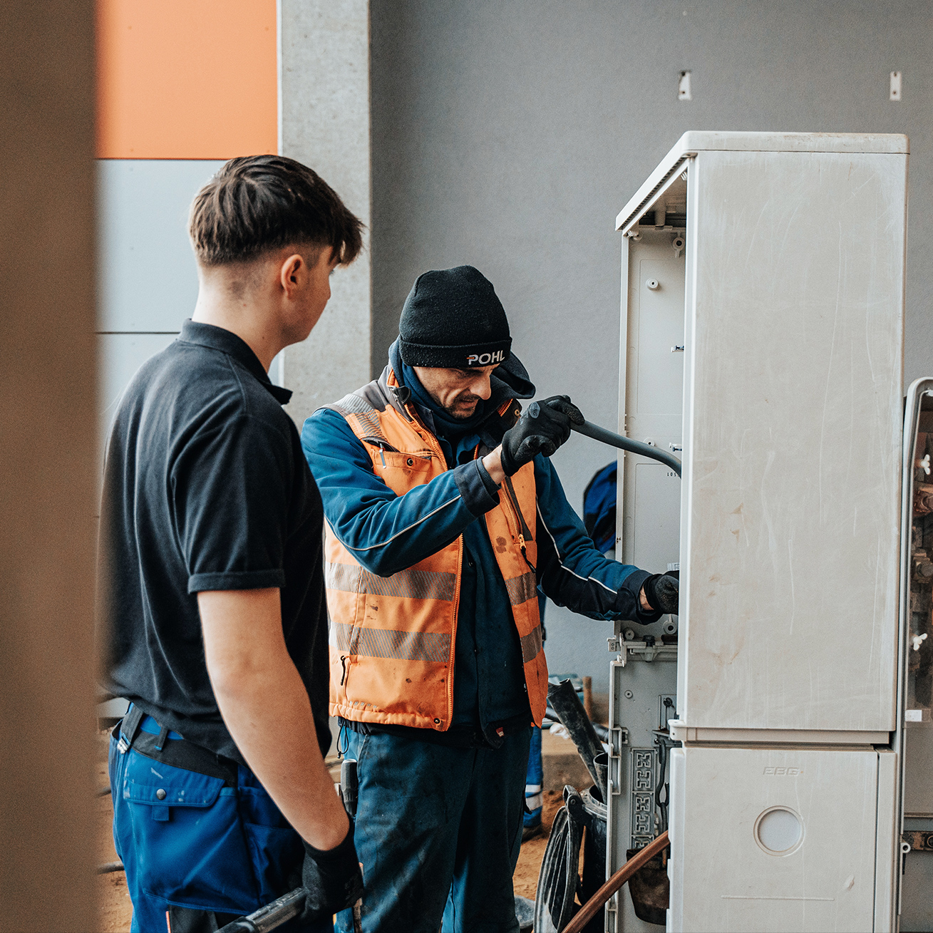 Links im Vordergrund steht eine Person im schwarzen Polo im Halbprofil; rechts daneben arbeitet ein Monteur in orangefarbener Warnweste und dunkelblauer Kleidung mit POHL-Mütze an einem geöffneten, grauen Straßen-Schaltschrank. Er hält mit behandschuhten Händen ein Rohr/Leerrohr und führt es in den Schrank ein; weitere geöffnete Fächer sind rechts sichtbar. Hintergrund: hellgraue Gebäudewand; Außenarbeitsplatz bei Tageslicht.