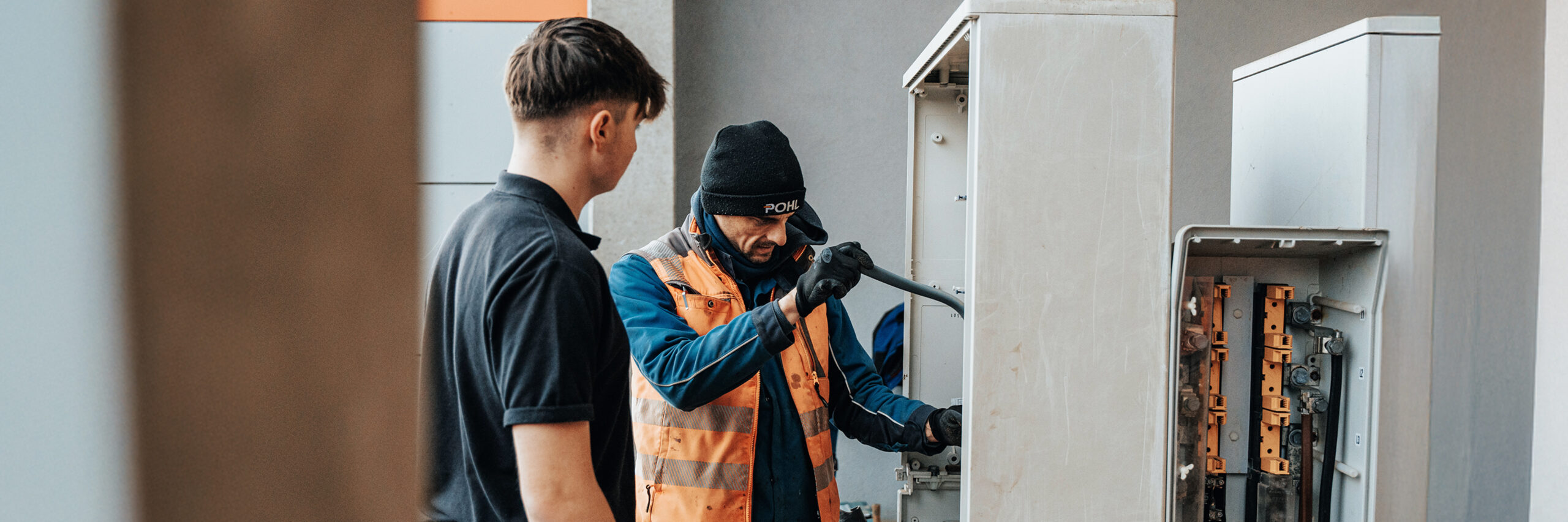 Links im Vordergrund steht eine Person im schwarzen Polo im Halbprofil; rechts daneben arbeitet ein Monteur in orangefarbener Warnweste und dunkelblauer Kleidung mit POHL-Mütze an einem geöffneten, grauen Straßen-Schaltschrank. Er hält mit behandschuhten Händen ein Rohr/Leerrohr und führt es in den Schrank ein; weitere geöffnete Fächer sind rechts sichtbar. Hintergrund: hellgraue Gebäudewand; Außenarbeitsplatz bei Tageslicht.