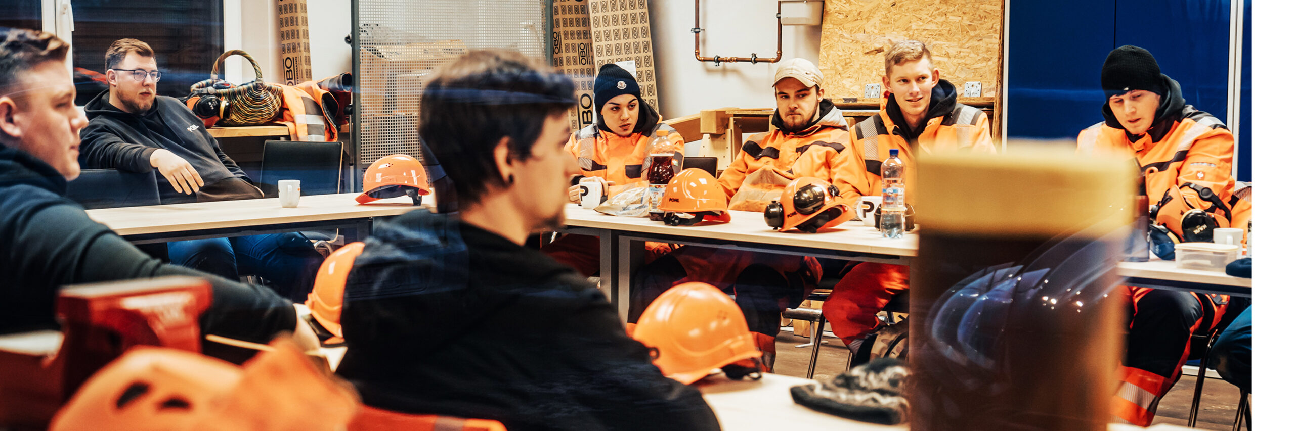 In der Mitte sitzen mehrere POHL-Mitarbeiter in orange Schutzkleidung an zusammengestellten Tischen. Vorne liegen orange Schutzhelme, Becher und Unterlagen; im Hintergrund stehen Regale und Material.