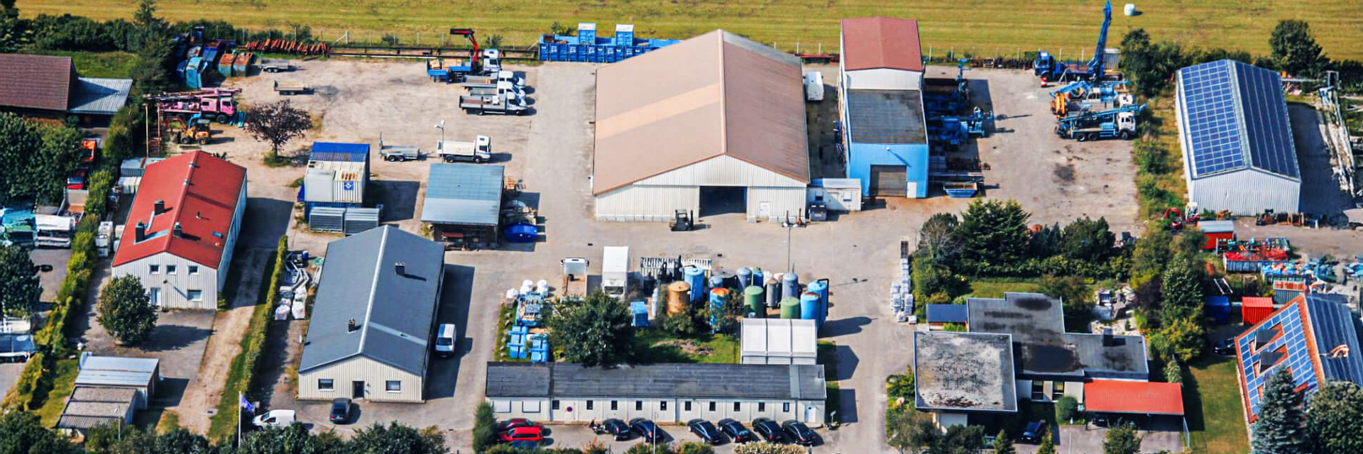 Standort Osterrönfeld: Vorne im Bild liegt ein rechteckiger Hof mit mehreren Hallen und Garagen. In der Mitte steht eine große Halle mit hellem Dach, links und rechts daneben weitere Hallen. Unten am Rand parken Autos und stehen Paletten sowie Fässer. Im Hintergrund grenzen Wiesen an das Gelände.
