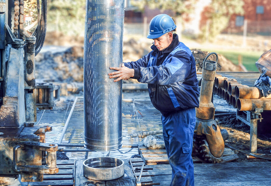 Ein Mann in blauer Arbeitskleidung und Schutzhelm steht auf einer Baustelle und hält ein großes, senkrecht stehendes Metallrohr fest. Das Rohr ist silbrig glänzend und etwa so breit wie sein Oberkörper. Er richtet es über einer runden Öffnung aus, die im Boden einer Gitterplattform eingelassen ist. Hinter ihm liegen weitere Rohre und Baumaschinenteile; die Umgebung ist staubig und wirkt wie ein aktives Bau- oder Bohrfeld. Die Szene ist in warmes Sonnenlicht getaucht, das die Metalloberflächen reflektieren lässt.