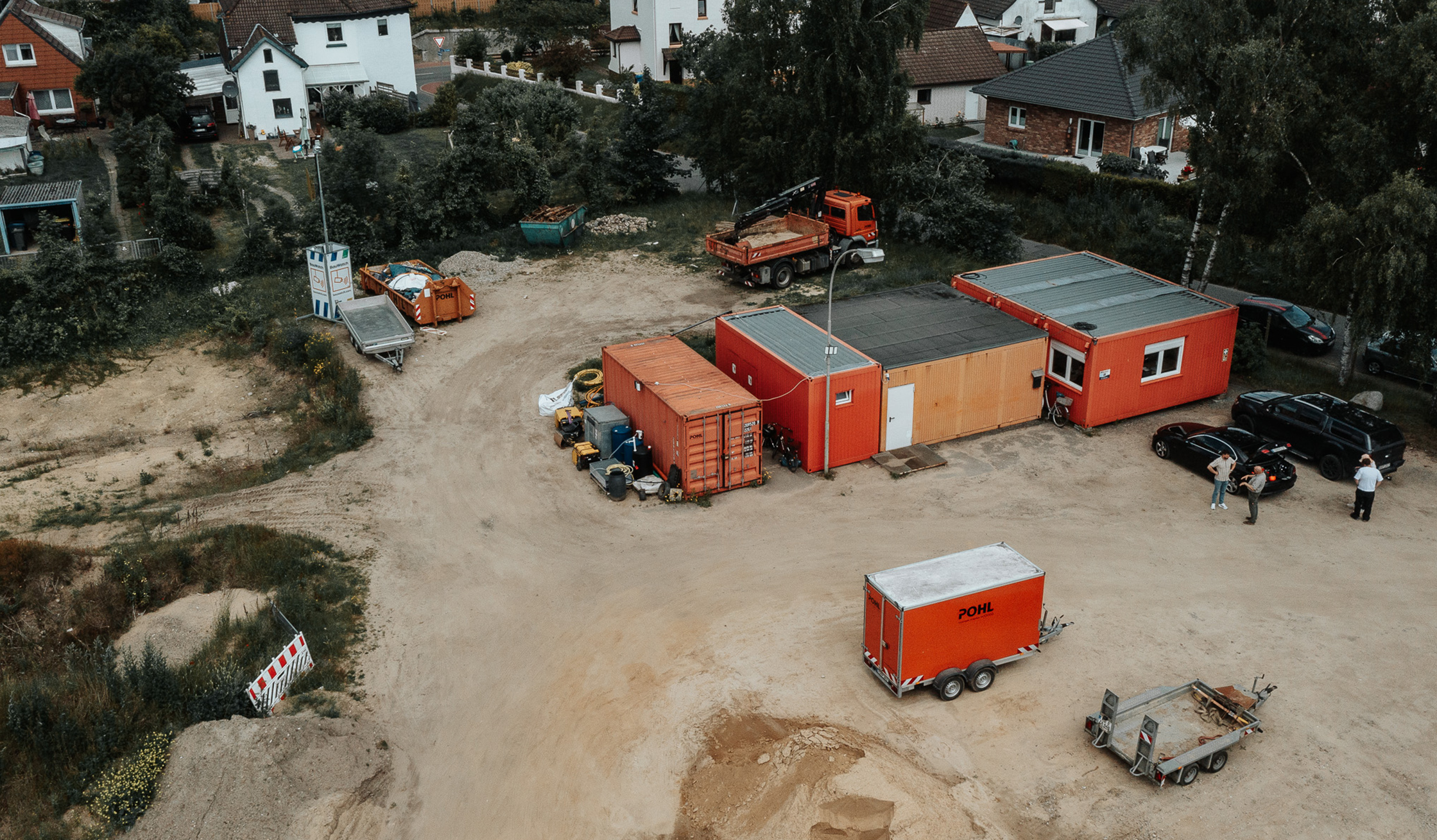 Standort Heide: Vorne im Bild steht ein offener Platz mit einem einzelnen POHL-Anhänger in Orange. In der Mitte gruppieren sich drei Container zu einem kleinen Bauzwischenlager; rechts daneben parken zwei dunkle Autos. Im Hintergrund stehen Wohnhäuser mit Gärten.