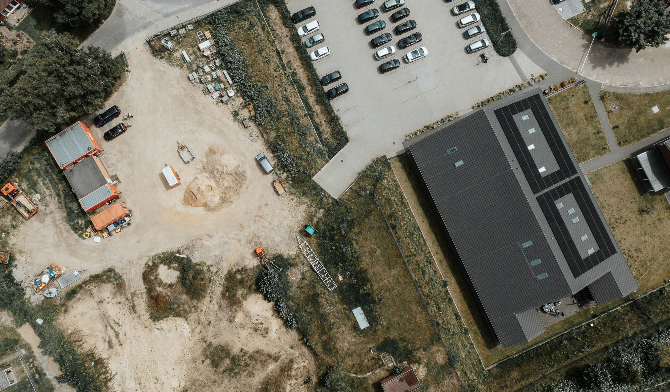Standort Heide: Oben rechts im Bild liegt ein großes Hallengebäude mit dunklem Dach und vielen Dachfeldern. Links daneben erstreckt sich ein grauer Parkplatz mit geordneten Autos. Unten links stehen mehrere Container und Materialstapel. Unten im Bild liegt eine helle Erdfläche mit Spuren.
