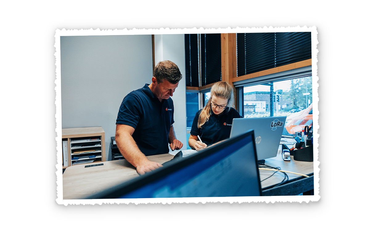 In der Mitte stehen zwei Personen an einem Schreibtisch und schauen auf Unterlagen. Rechts im Bild steht ein aufgeklappter Laptop vor einem Fenster mit Blick auf eine Straße. Links im Bild ist der unscharfe Rand eines Monitors zu sehen. Im Hintergrund steht ein Regal mit Fächern und Ordnern. Vorne im Bild liegen Kabel und Stifte auf der Tischplatte.