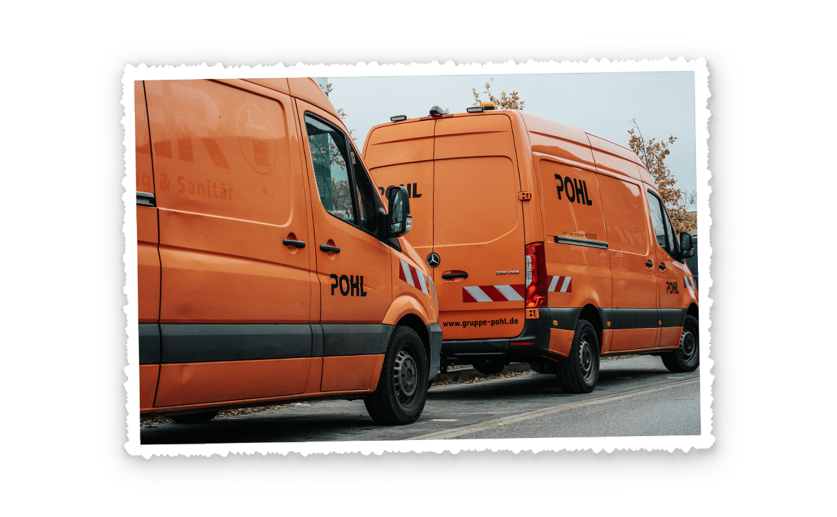 In der Mitte parken zwei orangefarbene POHL-Fahrzeuge hintereinander am Straßenrand. Links im Bild ist die Seitenwand des vorderen Fahrzeugs mit POHL-Schriftzug und weißen Warnstreifen zu sehen. Rechts im Bild steht der hintere Transporter mit großem POHL-Schriftzug auf der Hecktür. Im Hintergrund stehen helle Bäume mit dünnen Ästen.