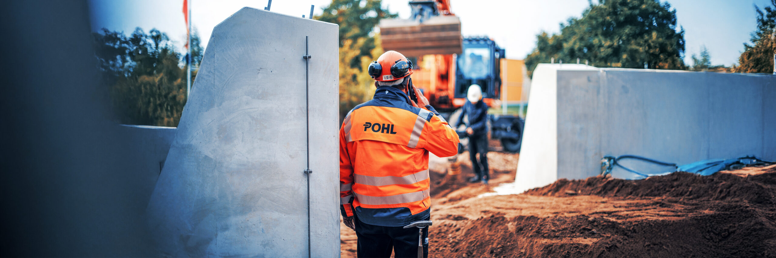 Mitarbeiter von POHL in orangener Schutzkleidung telefoniert auf einer Baustelle vor einem Bagger.