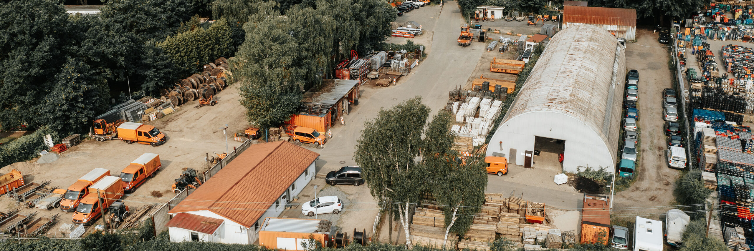 Standort Hamburg Hogenfeldweg: Vorne im Bild liegt ein Betriebshof mit vielen Materialstapeln. In der Mitte stehen mehrere POHL-Fahrzeuge in Orange sowie eine lange, helle Bogenhalle mit offenem Tor. Links am Rand grenzen hohe Bäume den Hof ab. Rechts im Bild stehen weitere Palettenreihen und geparkte Autos.