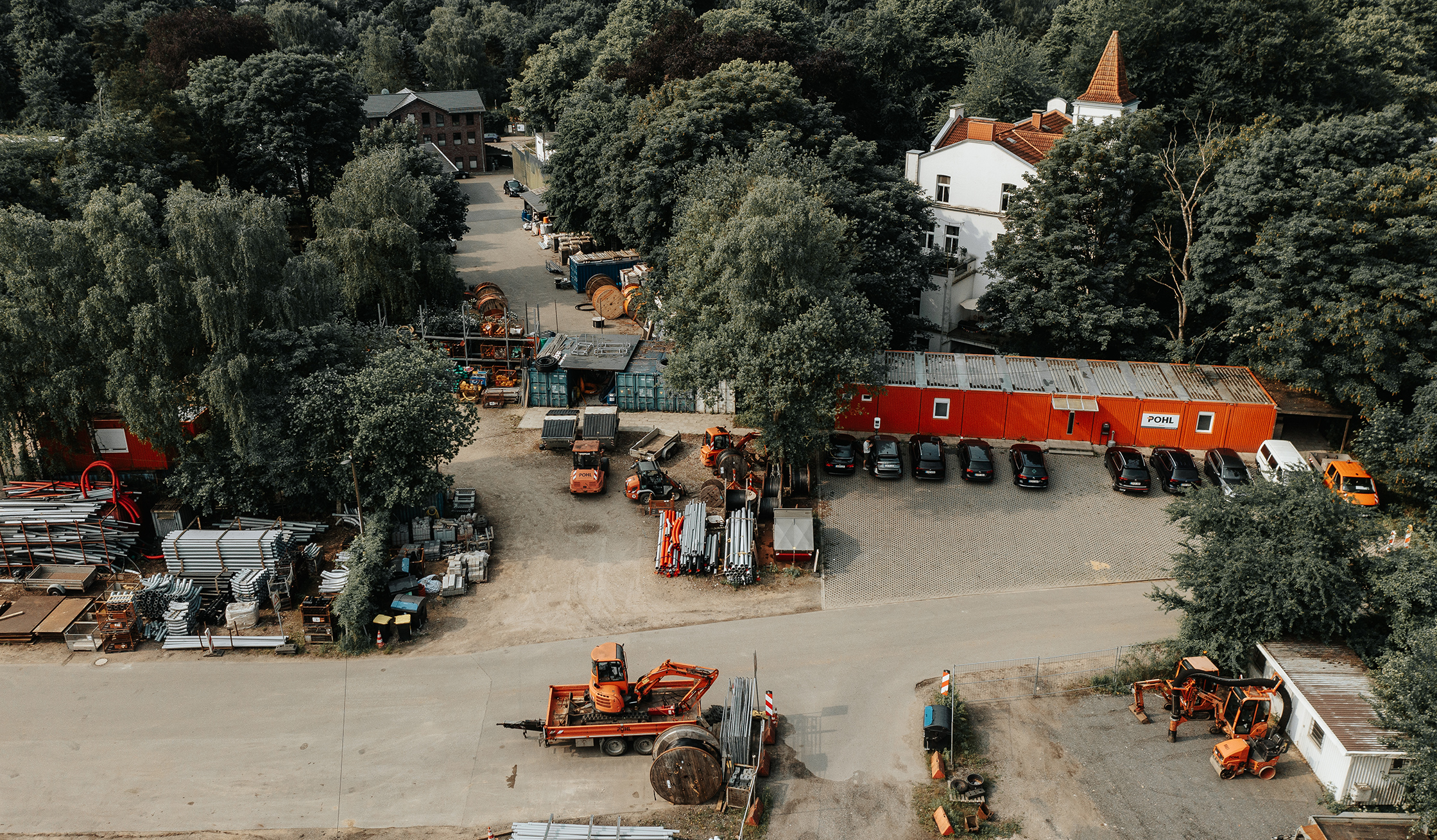 Standort Hamburg Hogenfeldweg: Vorne im Bild steht ein Hof mit Materialstapeln und mehreren orangefarbenen Maschinen. In der Mitte verläuft eine Reihe roter Container; an einem Container hängt ein Schild mit „POHL“. Rechts davor parken dunkle Autos. Im Hintergrund stehen viele Bäume und helle Gebäude.