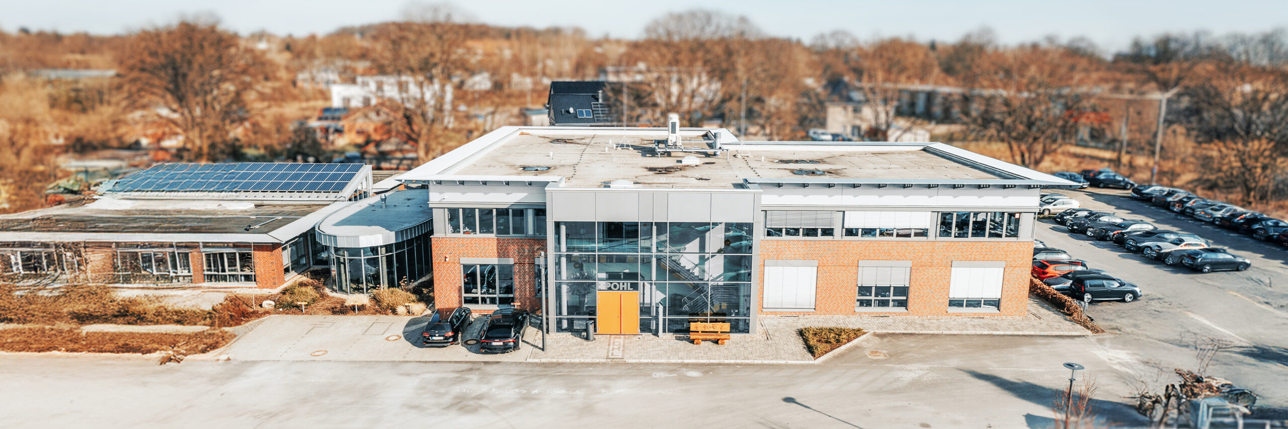 Standort Hohenwestedt: In der Mitte steht ein modernes Gebäude mit großem gläsernem Eingangsbereich; am Eingang ist der POHL-Schriftzug sichtbar. Vorne im Bild parken mehrere Pkw, rechts hinten steht ein langer Parkplatz mit weiteren Autos. Links schließt sich ein Gebäudeteil mit vielen Fenstern an.