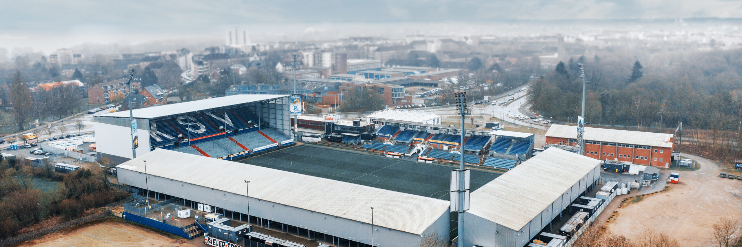 Referenz Erschließung Holsteinstadion Kiel: Luftaufnahme eines Fußballstadions: in der Mitte das Spielfeld, links eine hohe Tribüne mit großen weißen KSV-Buchstaben; rundherum Gebäude und Parkflächen.