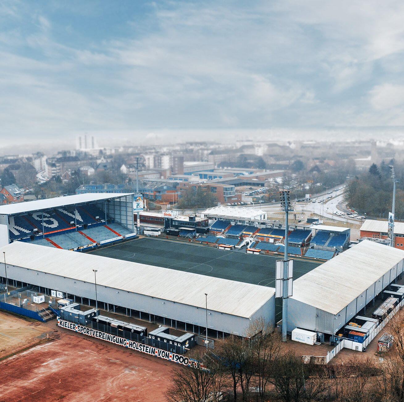 Referenz Erschließung Holsteinstadion Kiel: Luftaufnahme eines Fußballstadions: in der Mitte das Spielfeld, links eine hohe Tribüne mit großen weißen KSV-Buchstaben; rundherum Gebäude und Parkflächen.