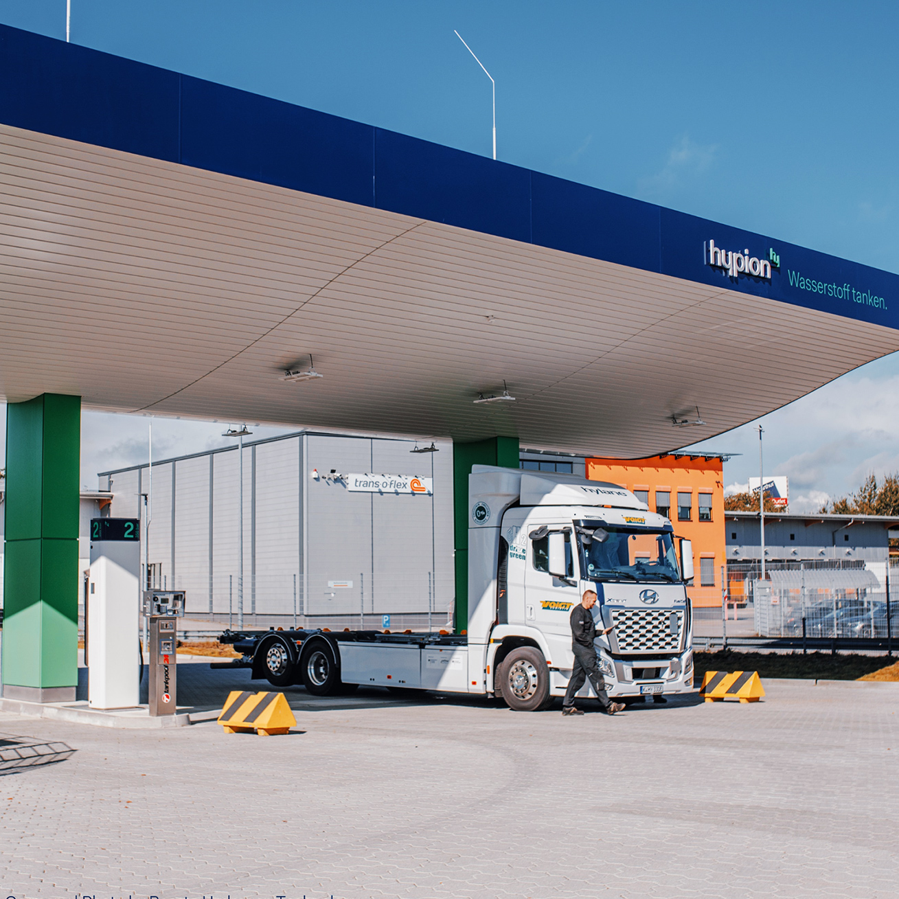 Referenz Hypion Wasserstofftankstelle Neumünster: In der Mitte steht eine überdachte Wasserstoff-Tankstelle mit blauem Dach und grünen Pfeilern. Rechts im Bild hält ein weißer Lastwagen unter dem Dach; ein Mann geht davor vorbei. Im Hintergrund stehen Hallengebäude hinter einem Zaun.