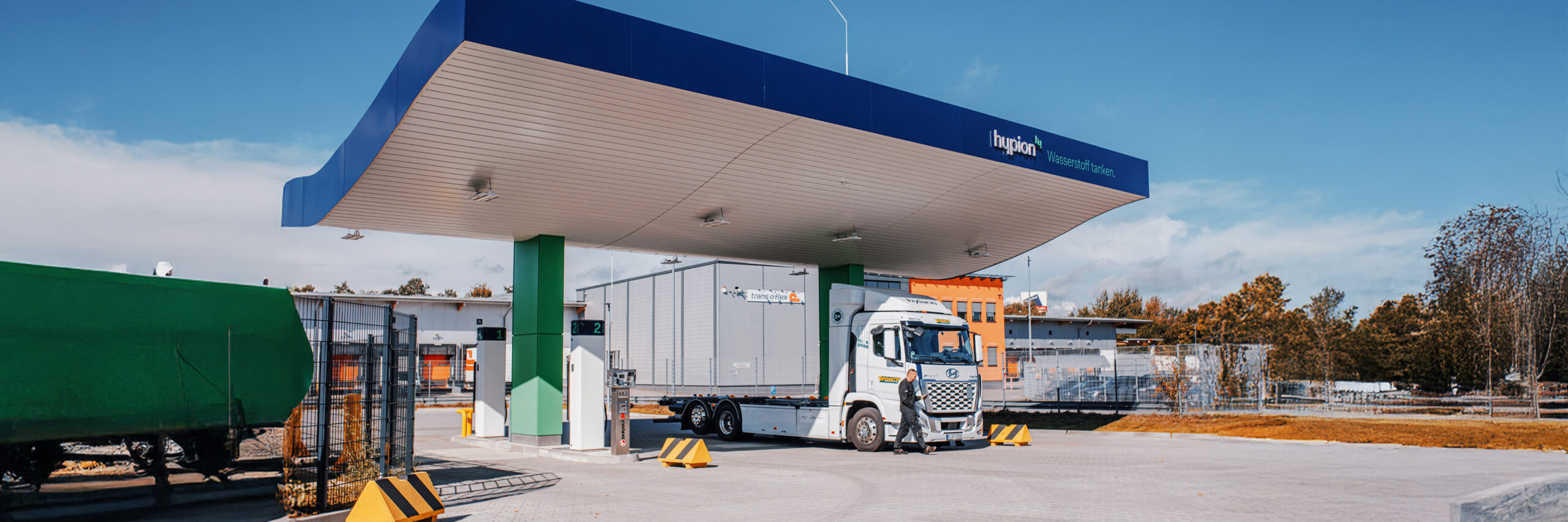 Referenz Hypion Wasserstofftankstelle Neumünster: In der Mitte steht eine überdachte Wasserstoff-Tankstelle mit blauem Dach und grünen Pfeilern. Rechts im Bild hält ein weißer Lastwagen unter dem Dach; ein Mann geht davor vorbei. Im Hintergrund stehen Hallengebäude hinter einem Zaun.