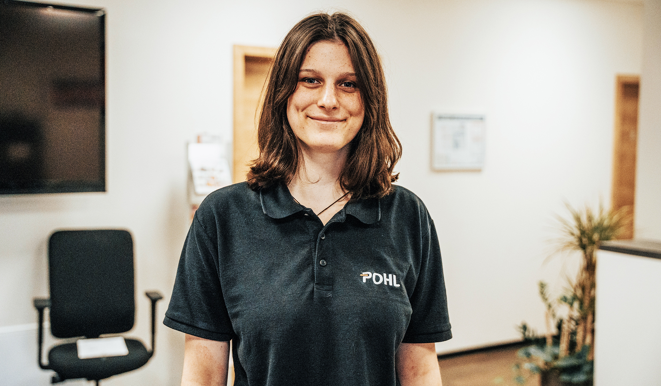 In der Mitte steht eine Person im schwarzen POHL-Polo an einer Theke und hält einen Stift über ein Formular. Links hinten steht ein Bürostuhl; rechts sind Schränke und eine Pflanze zu sehen.