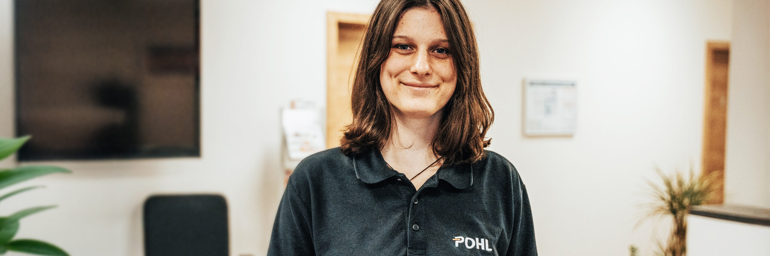 In der Mitte steht eine Person im schwarzen POHL-Polo an einer Theke und hält einen Stift über ein Formular. Links hinten steht ein Bürostuhl; rechts sind Schränke und eine Pflanze zu sehen.