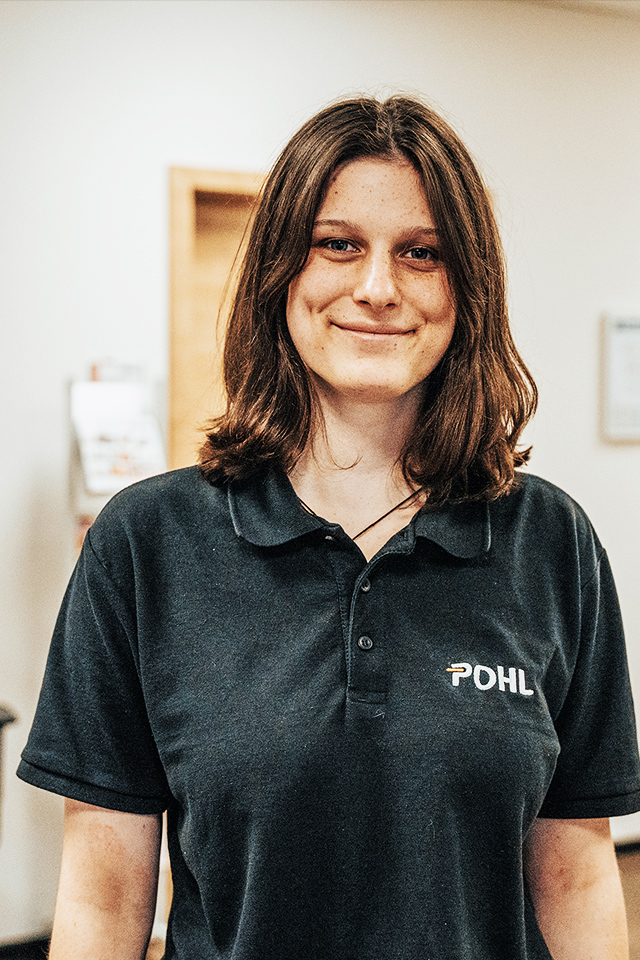 In der Mitte steht eine Person im schwarzen POHL-Polo an einer Theke und hält einen Stift über ein Formular. Links hinten steht ein Bürostuhl; rechts sind Schränke und eine Pflanze zu sehen.